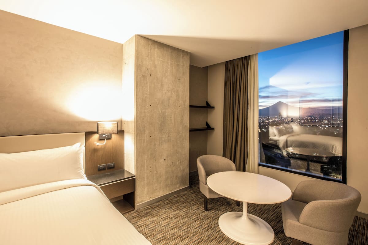 A hotel room with a bed, a small table, two chairs, a nightstand, a lamp, a window with a city and mountain view, and a concrete accent wall.