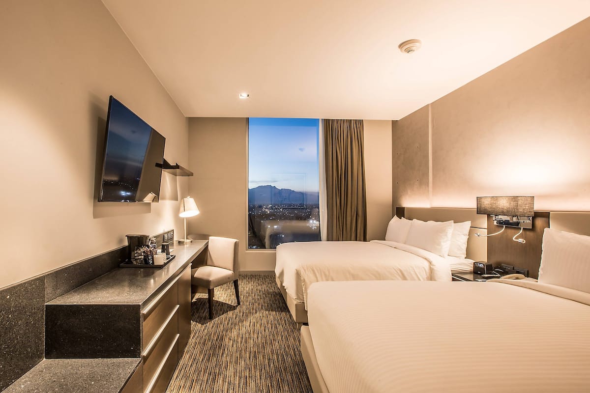 A hotel room with two beds, a desk, a chair, a large window with a city view, and a mounted TV.