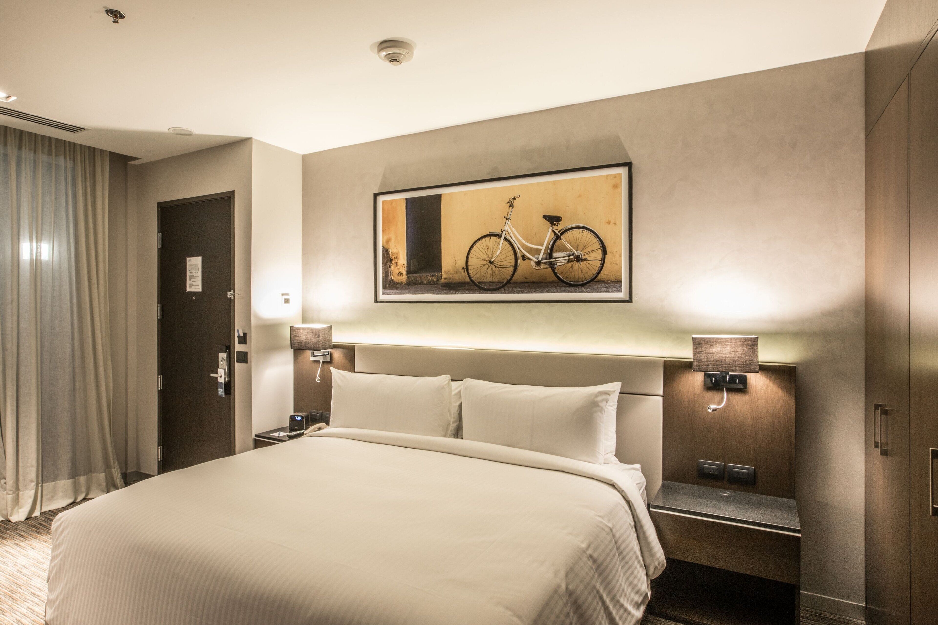 A hotel room with a large bed, a headboard with a mounted picture, bedside lamps, a nightstand, and a door.