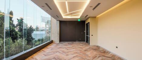 A modern hotel corridor with a herringbone-patterned floor, large windows offering a view of greenery, and a ceiling with linear lighting fixtures.