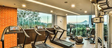 Un gimnasio con cintas de correr, máquinas de pesas y un amplio ventanal que ofrece vistas a la vegetación y una montaña.