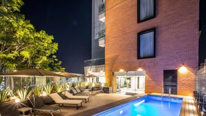 Un área de piscina en la azotea con sillones de descanso y sombrillas, rodeada por un edificio de ladrillo con ventanas grandes.
