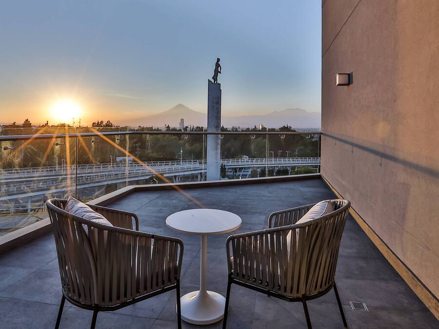 A balcony with two chairs and a small table, overlooking a cityscape with a statue and mountains in the background.