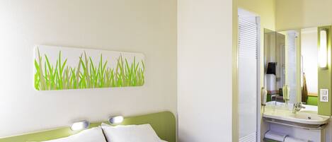 Una habitación de hotel con cama, lámparas de noche, cabecera verde, un cuadro en la pared que representa hierba verde y un baño con lavamanos y espejo.