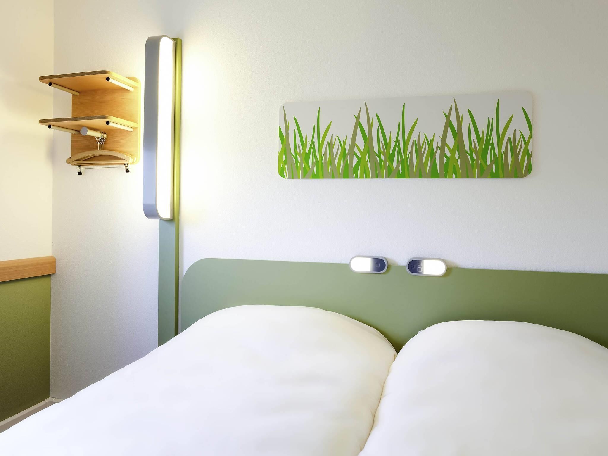Ein Bett mit zwei Kissen, einer Wandleuchte und einem Kunstwerk mit Grasmuster.