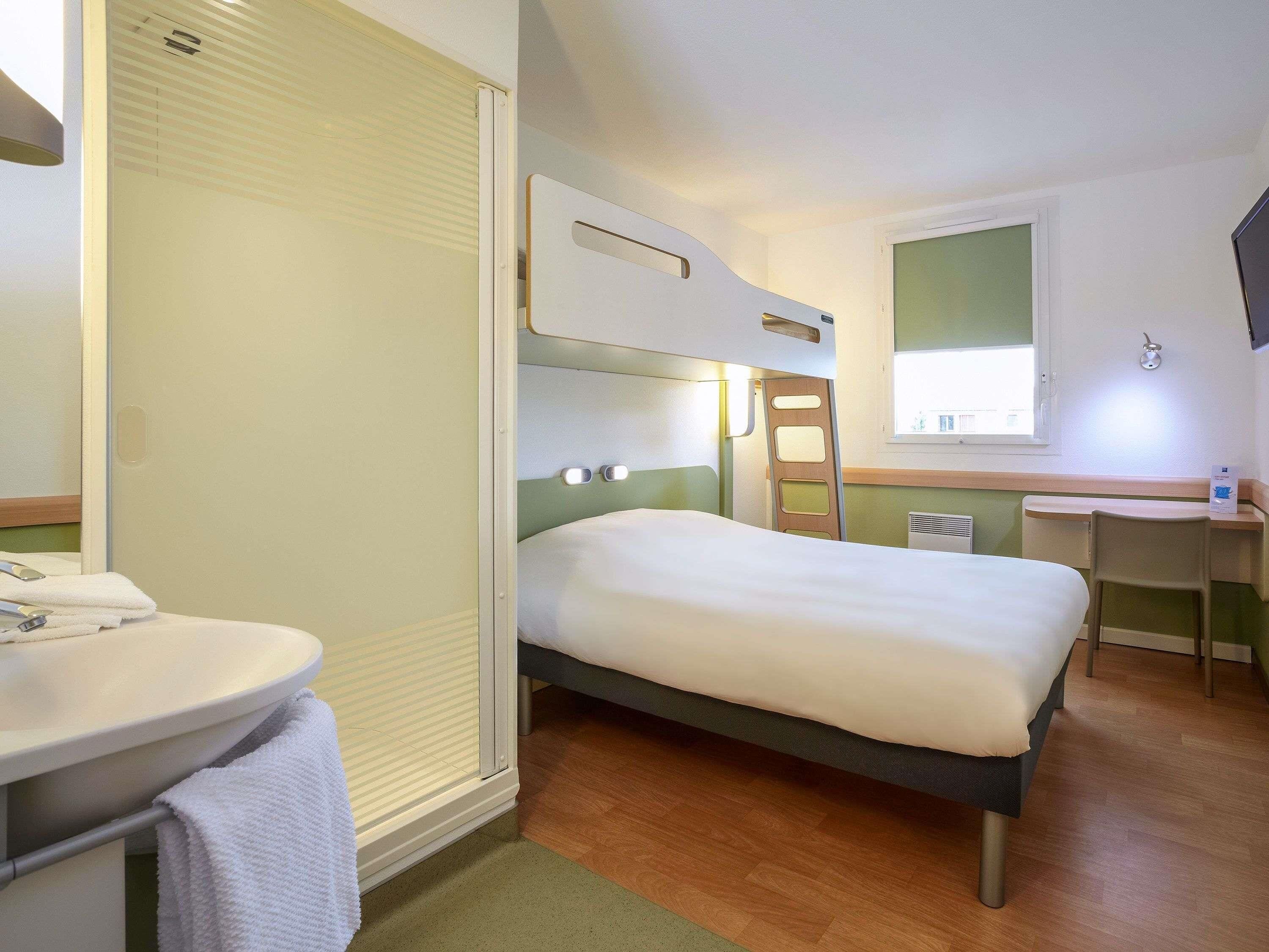 Habitación de hotel pequeña con una cama individual, un escritorio, una silla, una mesita de noche y un baño con inodoro y lavamanos.