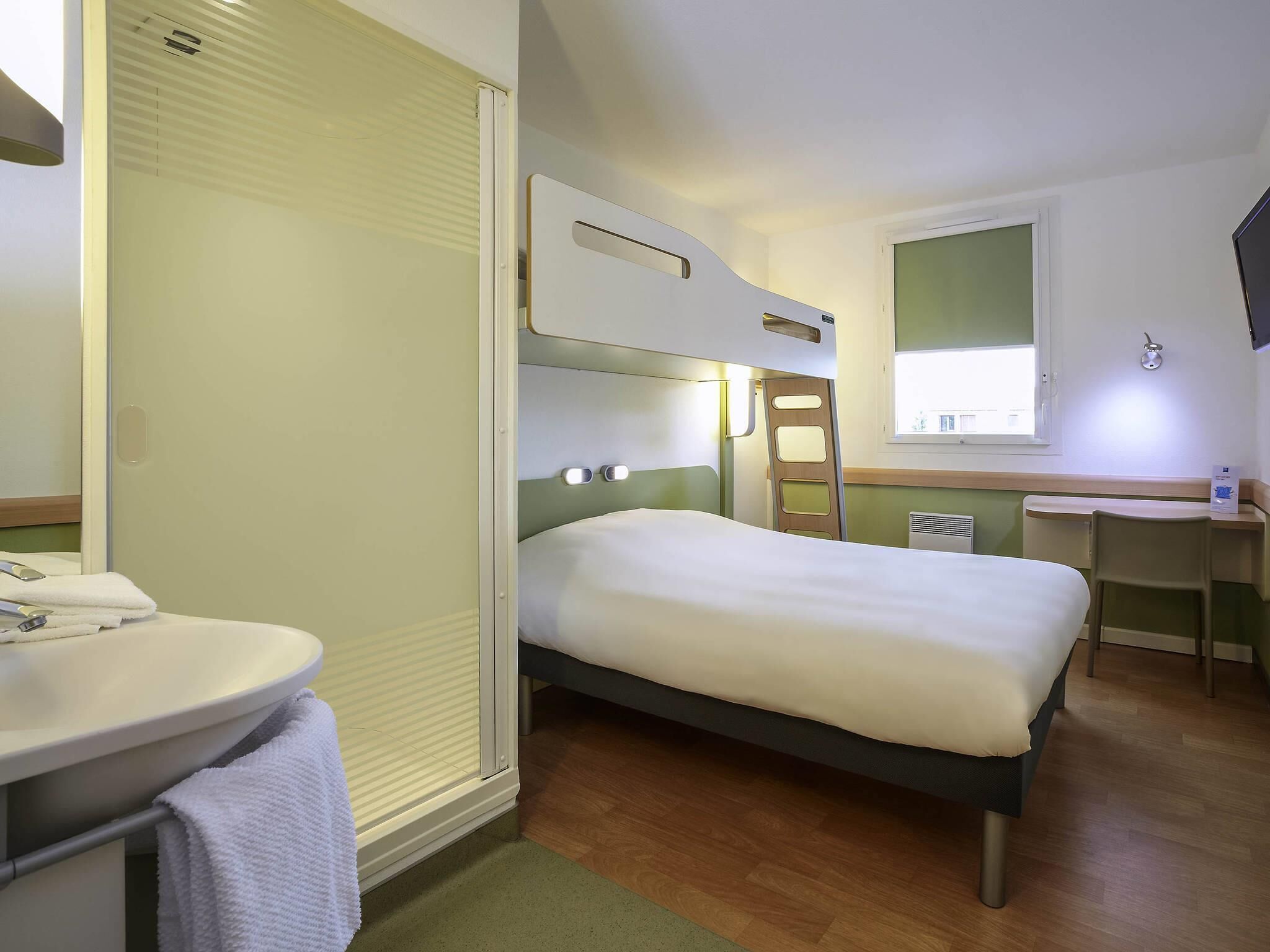 Ein kleines Hotelzimmer mit einem Einzelbett, einem kleinen Schreibtisch, einem Badezimmer mit Waschbecken und Spiegel sowie einem Fenster mit Tageslicht.