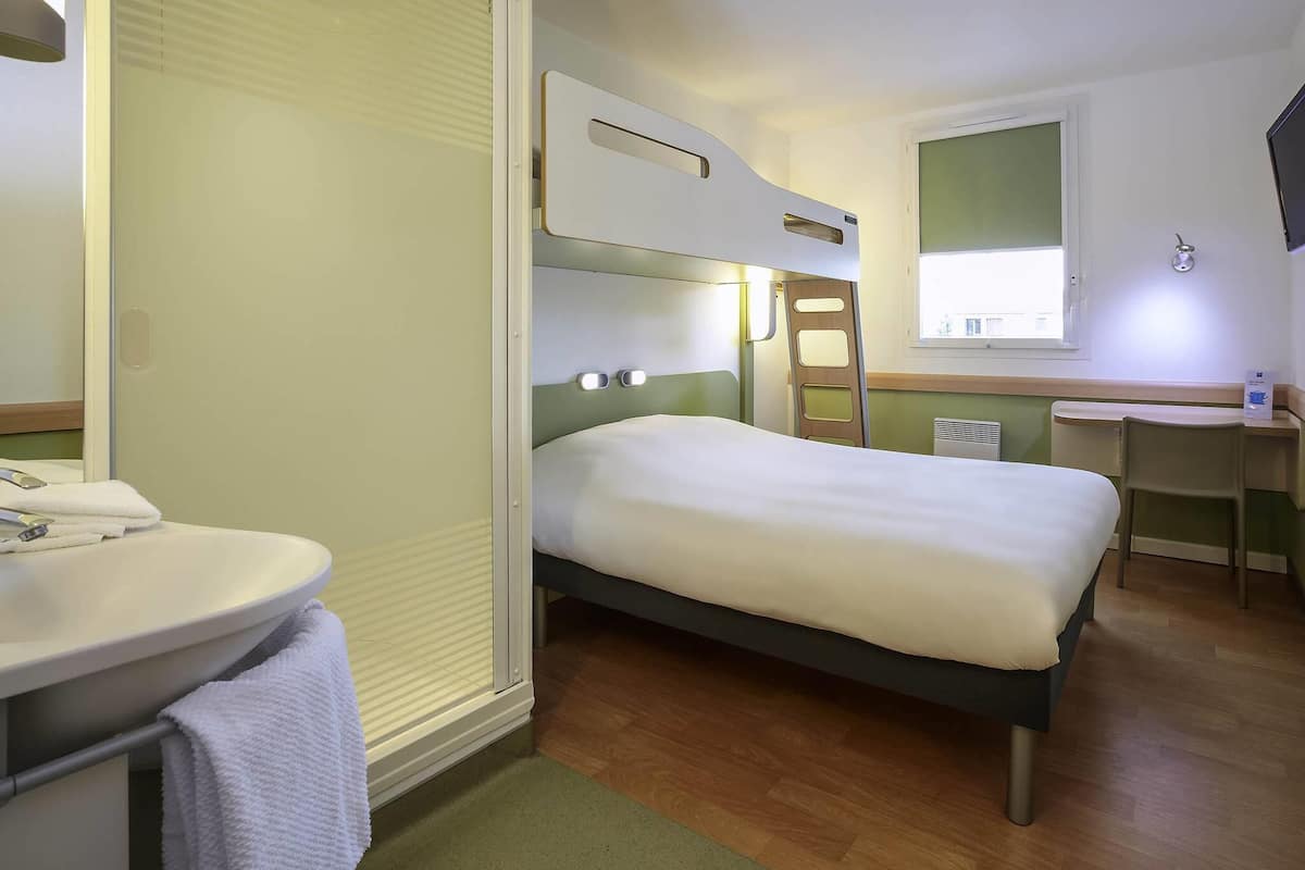 Ein kleines Hotelzimmer mit einem Einzelbett, einem kleinen Schreibtisch, einem Badezimmer mit Waschbecken und Spiegel sowie einem Fenster mit Tageslicht.