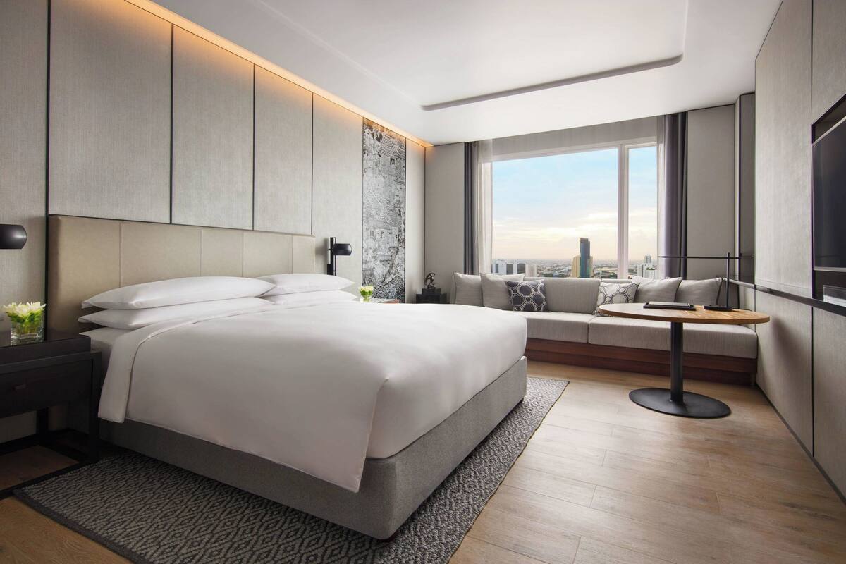 A modern hotel room with a large bed, a seating area with a table, and a city view through large windows.
