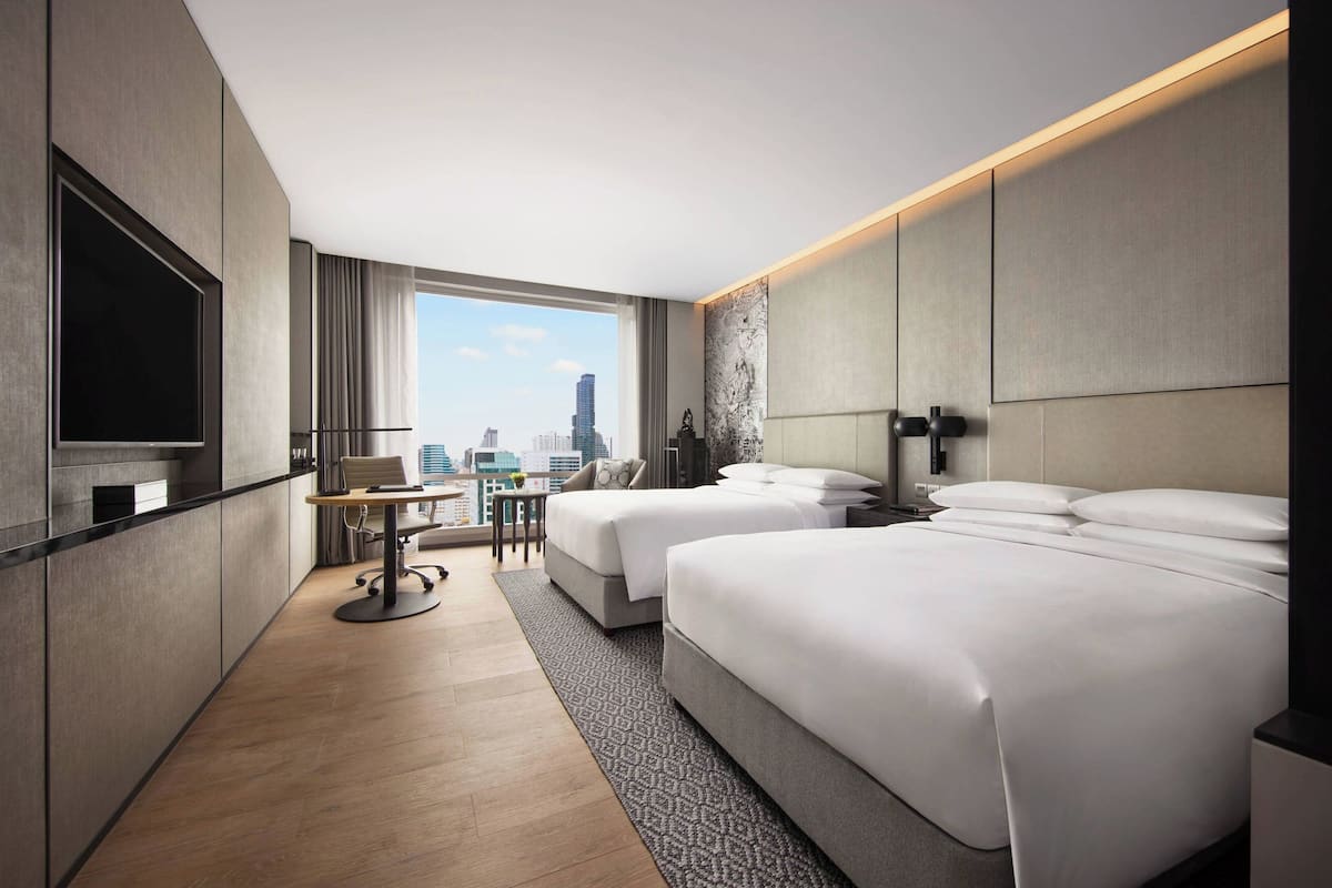 A modern hotel room with two beds, a desk, a chair, a television, and a city view.