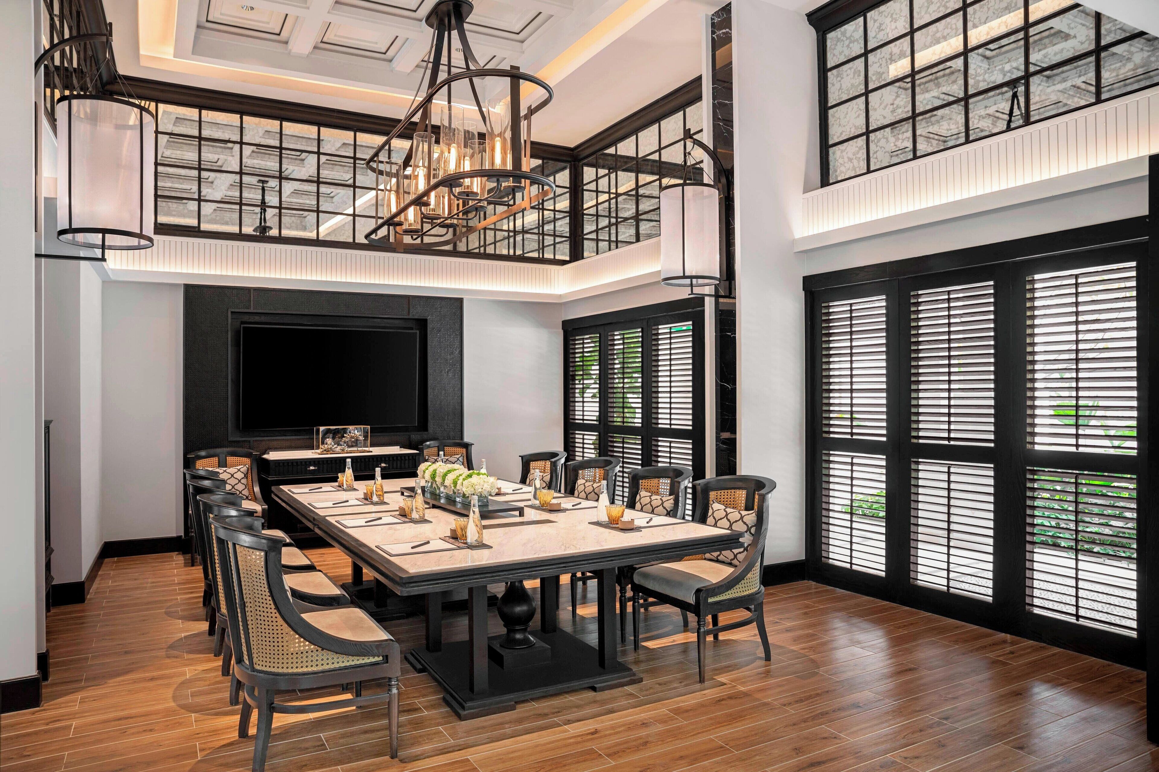 Uma ampla sala de jantar com uma mesa grande, várias cadeiras, um candelabro e janelas grandes com persianas.