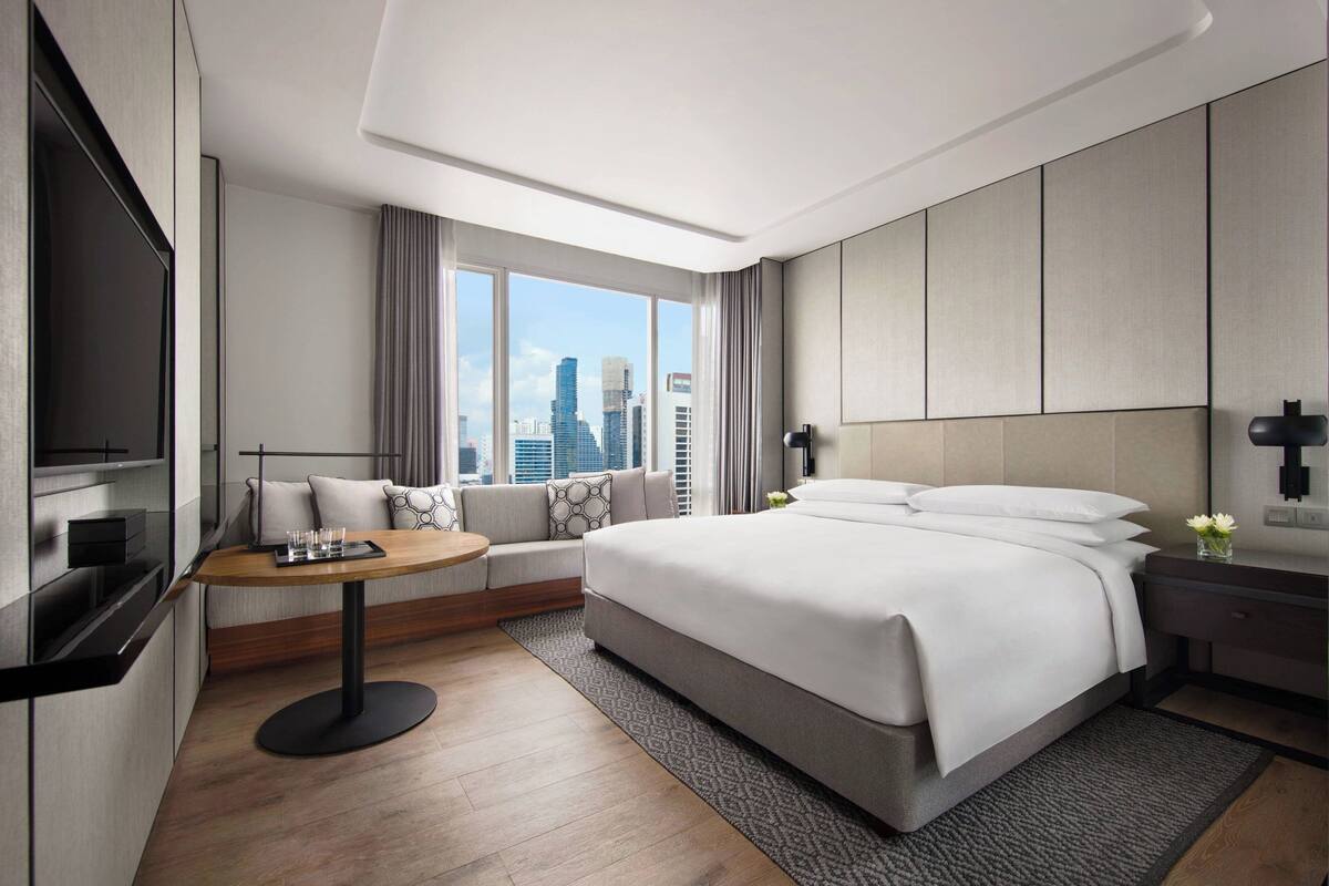 A modern hotel room with a large bed, a seating area with a table, a TV, and a city view.