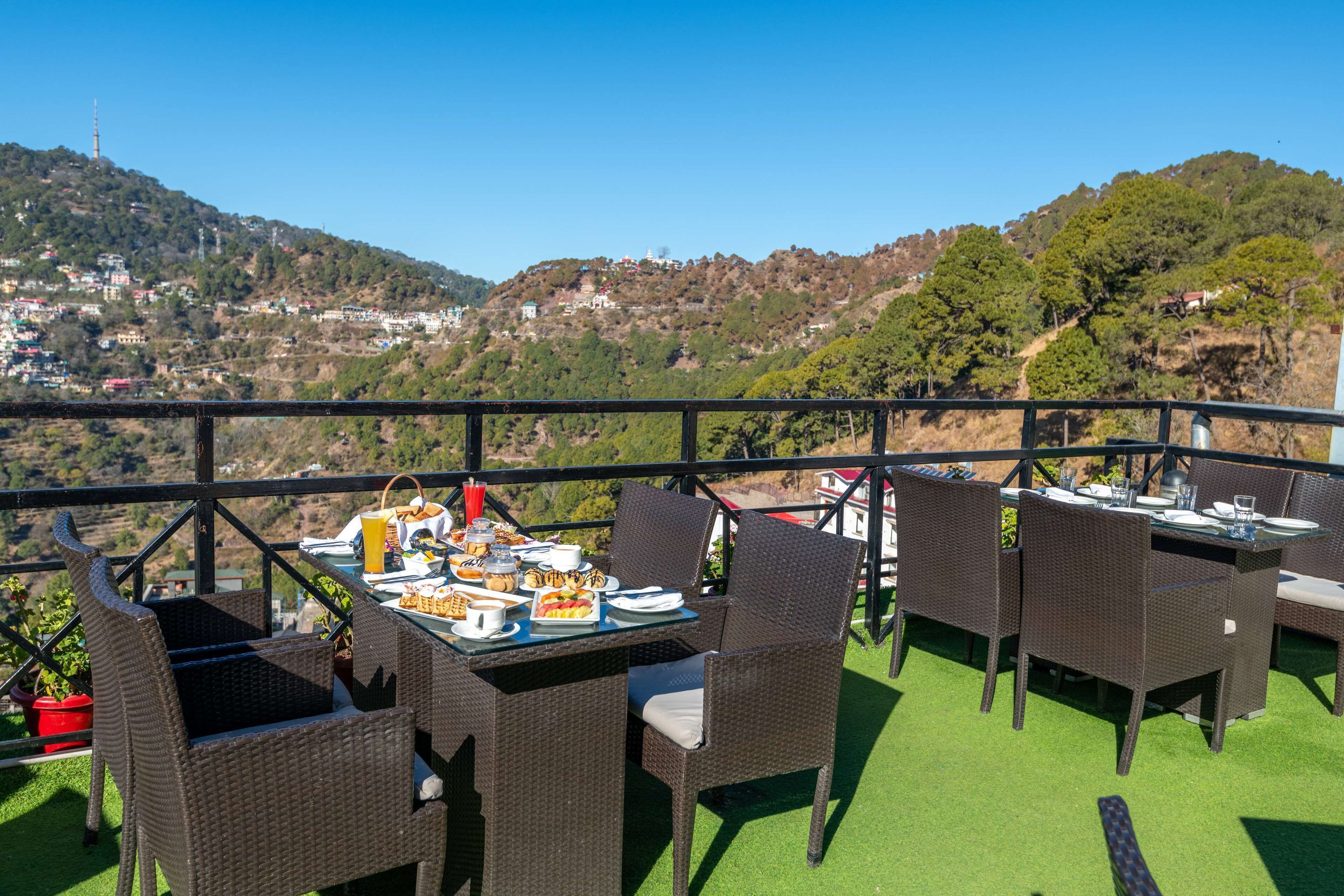 Un área de comedor al aire libre con mobiliario de mimbre y una mesa preparada para una comida, con vistas a un paisaje montañoso con edificios.