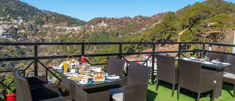 Un área de comedor al aire libre con mobiliario de mimbre y una mesa preparada para una comida, con vistas a un paisaje montañoso con edificios.