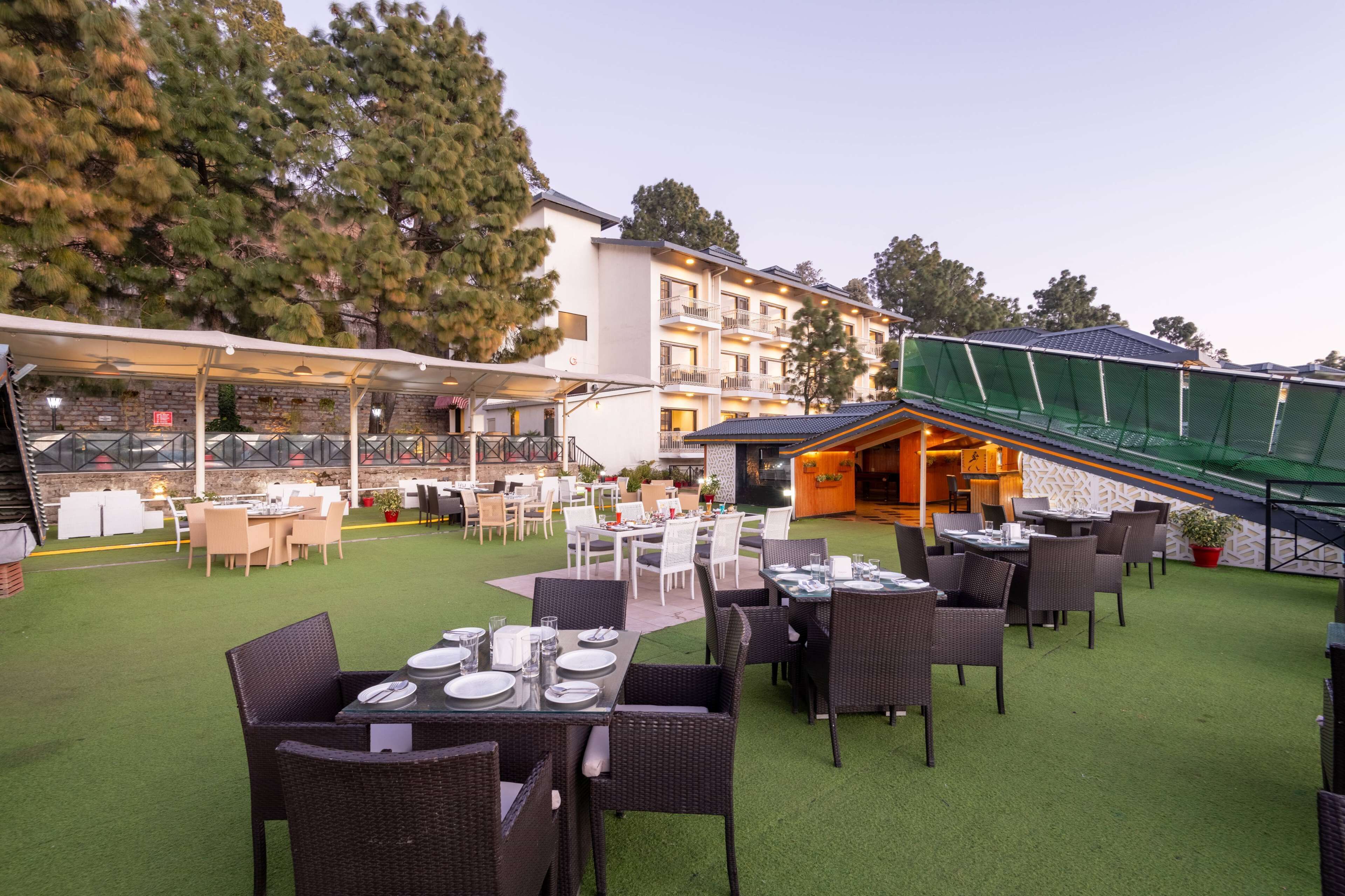 Un área de comedor al aire libre con mesas y sillas, un espacio de estar cubierto y un edificio al fondo.