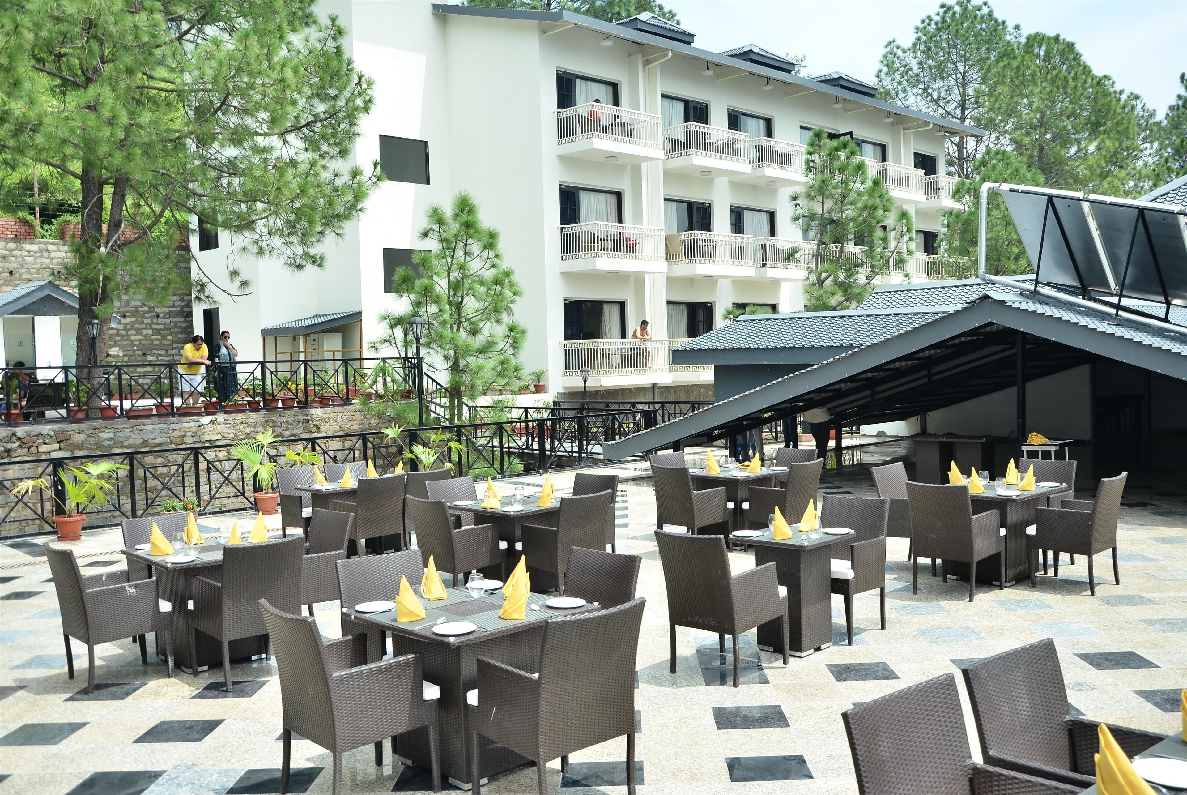 An outdoor dining area with wicker tables and chairs, potted plants, and a building with balconies in the background.