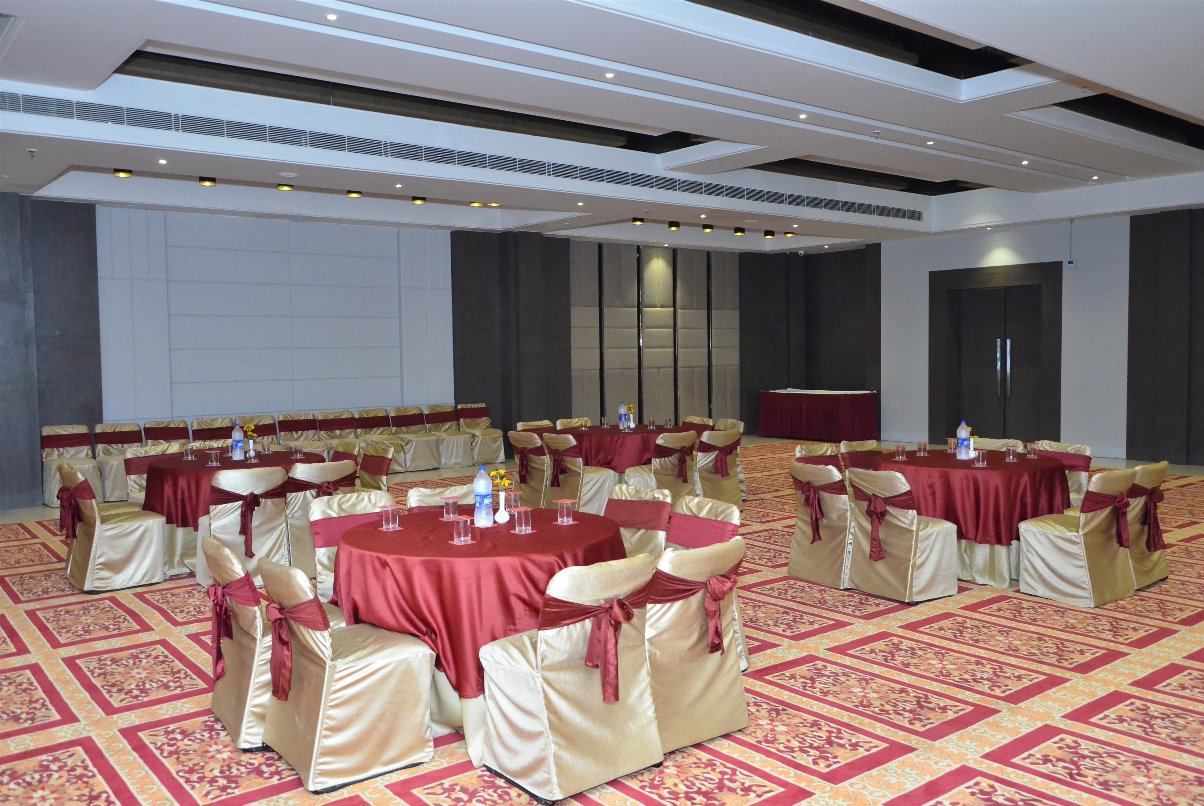 Een feestzaal met ronde tafels bedekt met rode tafelkleden, gerangschikt in rijen met stoelen bekleed met beige stof en rode sashes. De vloer heeft een kamerbreed tapijt, en de ruimte heeft een hoge plafond met inbouwverlichting.