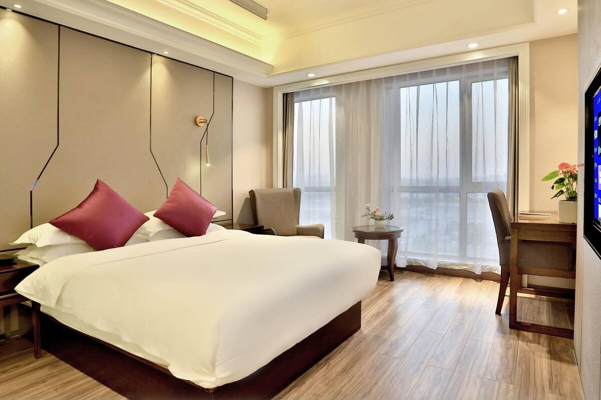 Ein Hotelzimmer mit einem großen Bett, einem Schreibtisch, einem Sessel, einem Fernseher und einem Fenster mit durchsichtigen Vorhängen.