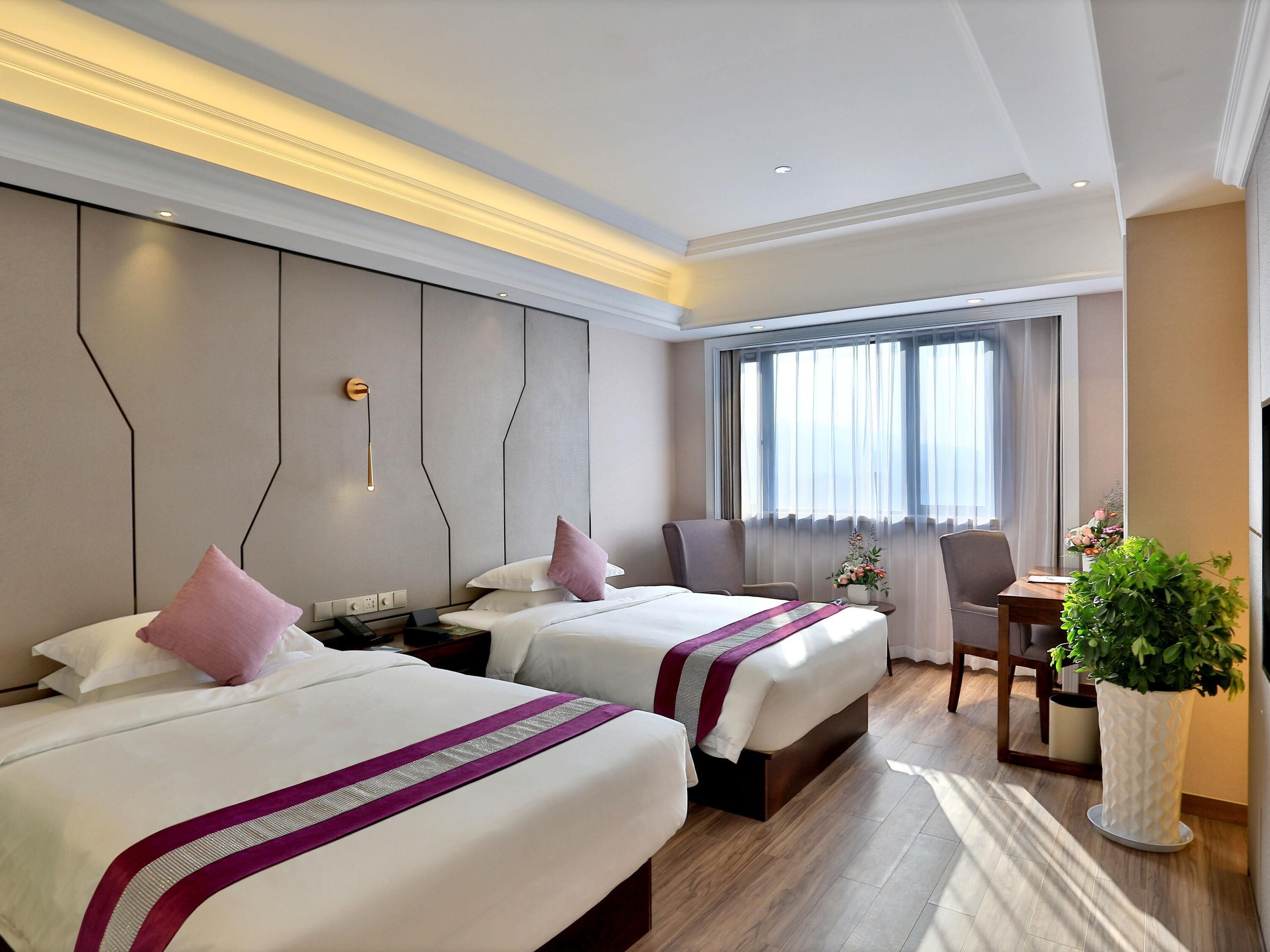 Une chambre d’hôtel avec deux lits, un bureau, une chaise, une plante en pot et une grande fenêtre avec des rideaux.