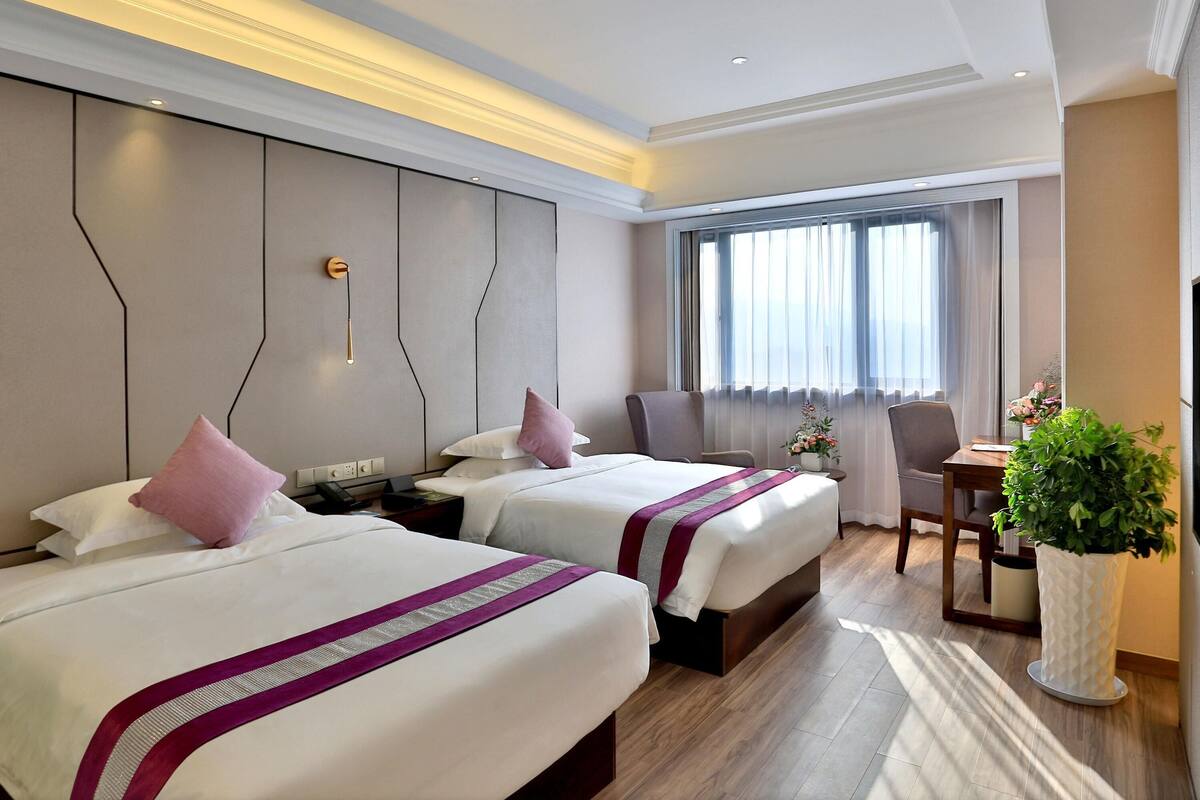 Ein Hotelzimmer mit zwei Betten, einem Schreibtisch, einem Stuhl, einer Zimmerpflanze und einem großen Fenster mit Vorhängen.