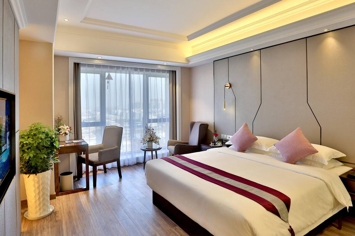 Ein Hotelzimmer mit einem großen Bett, einem Schreibtisch mit Stuhl, einem kleinen Tisch mit Vase und einem großen Fenster.