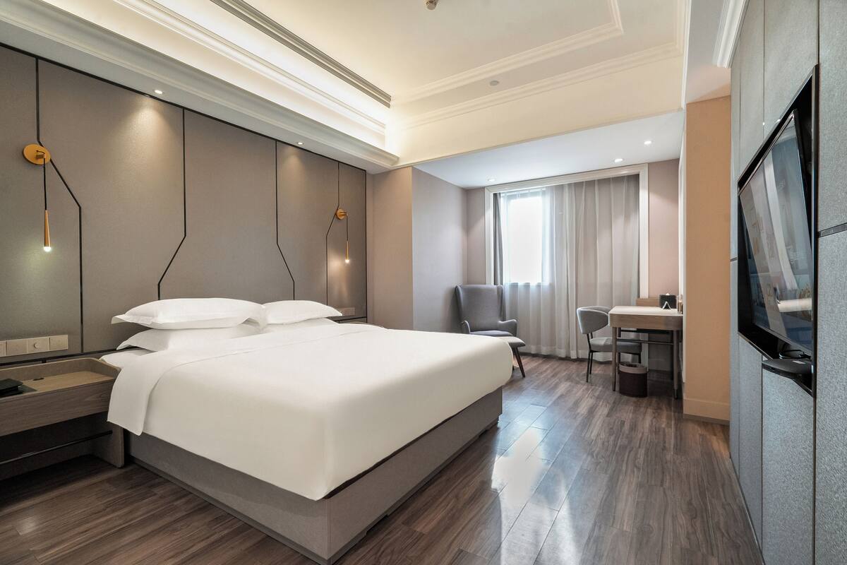 Ein modernes Hotelzimmer mit einem großen Bett, Nachttischen, einem Schreibtisch, einem Sessel, einem Fernseher und einem Fenster mit Vorhängen.