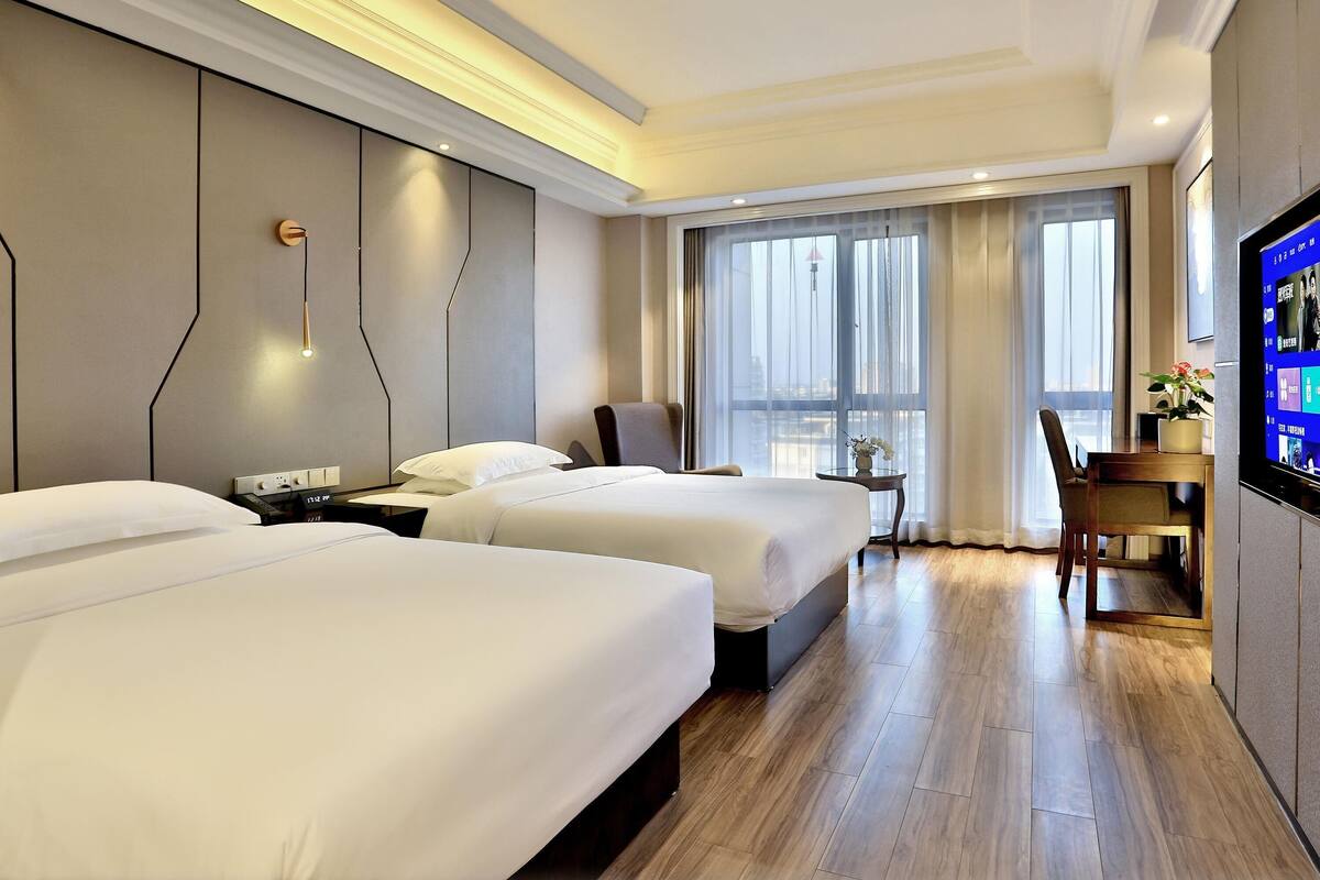 Ein Hotelzimmer mit zwei Betten, einem Schreibtisch, einem Stuhl, einem Fernseher und einem großen Fenster mit Vorhängen.