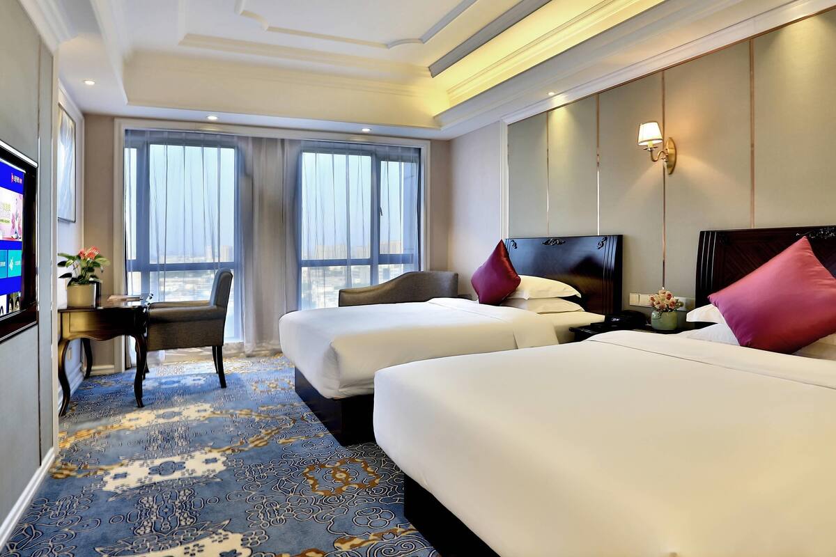 Ein Hotelzimmer mit zwei Betten, einem Fernseher, einem Schreibtisch mit Stuhl und großen Fenstern.