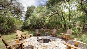 A circular stone fire pit surrounded by wooden Adirondack chairs in a forested area. -  Chic Hill Country Oasis (Driftwood)