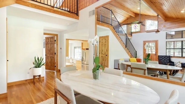 A spacious living area with a high ceiling, wooden beams, a dining table with chairs, and a staircase leading to an upper level. -  Chic Hill Country Oasis (Driftwood)