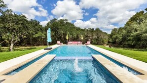A rectangular swimming pool with a water feature in the center, surrounded by a paved deck and green lawns. -  Chic Hill Country Oasis (Driftwood)