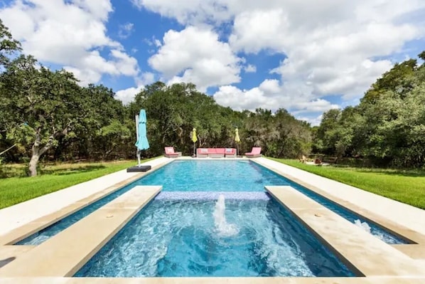 Una piscina rectangular con un elemento acuático en el centro, rodeada por un solárium empedrado y césped verde.