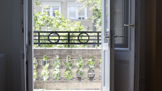 Een open deur die leidt naar een balkon met een ijzeren hekwerk en uitzicht op gebouwen en bomen.