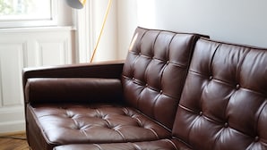 Een bruine leren bank met een gestuufde rugleuning in een kamer met een witte muur en een raam.