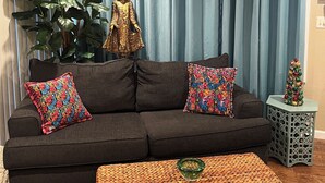 Een woonkamer met een donkere bank, kleurrijke kussens, een Boeddhabeeldje, een potplant, een kleine kerstboom en een geweven pouf.