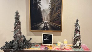 Een receptiegedeelte met een ingelijste foto van een weg omzoomd door bomen, een bord met de tekst welkom, twee kleine kerstboommetjes, een kaars en een decoratieve tafel.