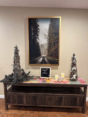 Un espace d’accueil avec une photographie encadrée, un panneau d’accueil, deux sapins de Noël et une petite table sur laquelle se trouvent une bougie et une tasse.