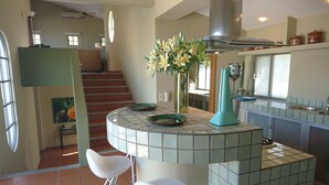Una cocina con una isla azulejada, dos taburetes blancos de bar, un jarrón con flores y una escalera que conduce a otra habitación.