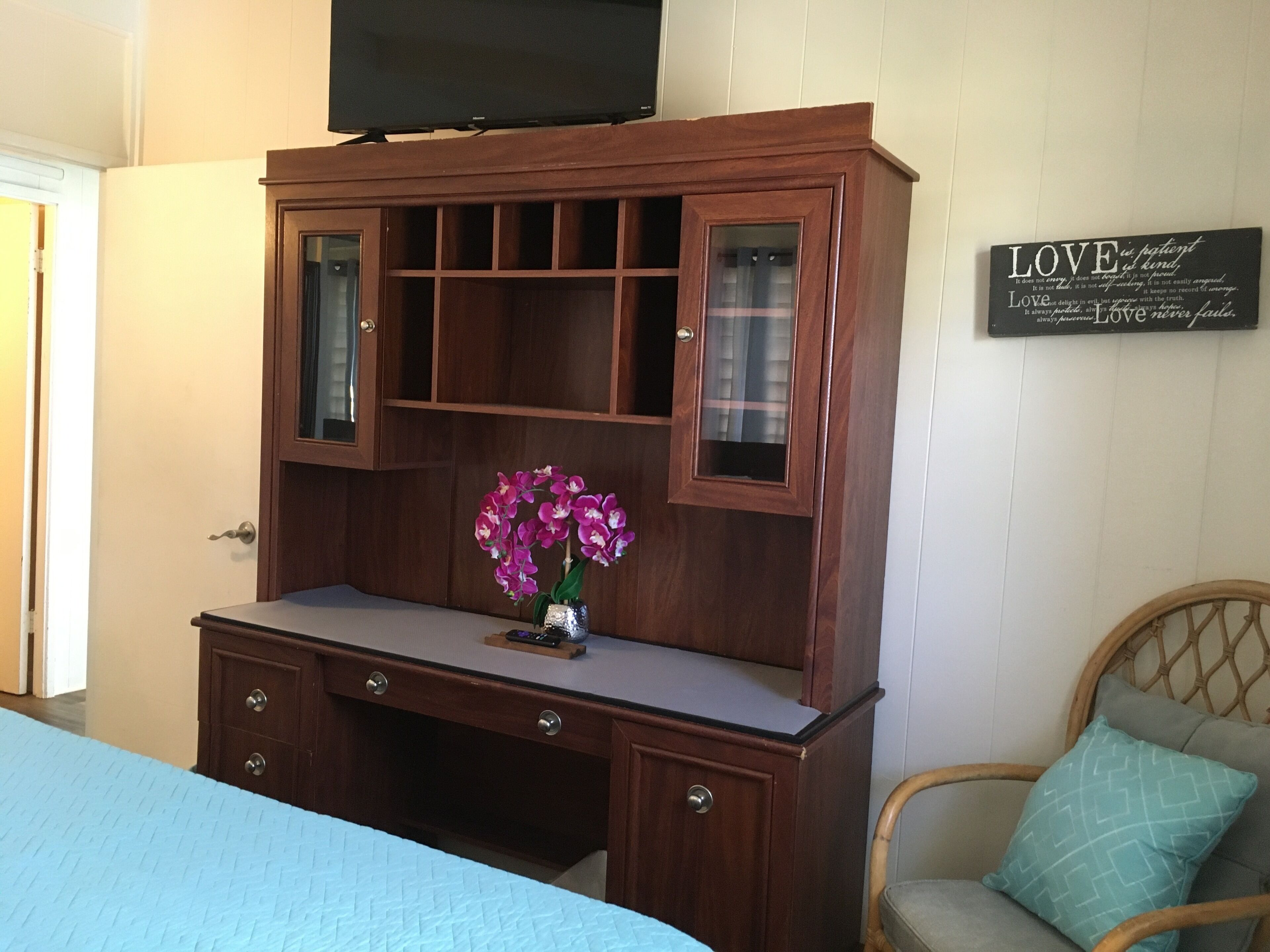 A bedroom with a wooden dresser, a television on top, a bed with a blue bedspread, and a chair with a cushion.