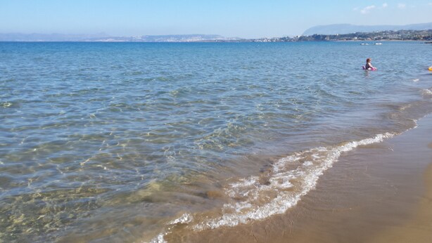 Una playa con agua cristalina, una persona en el agua y una costa lejana con edificios.