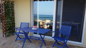 Un patio con una mesa y sillas azules, una planta decorativa y vista al mar a través de un ventanal.