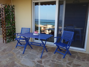 Eine Terrasse mit blauem Tisch und Stühlen, einer Zierpflanze und Blick auf das Meer durch eine Glastür.