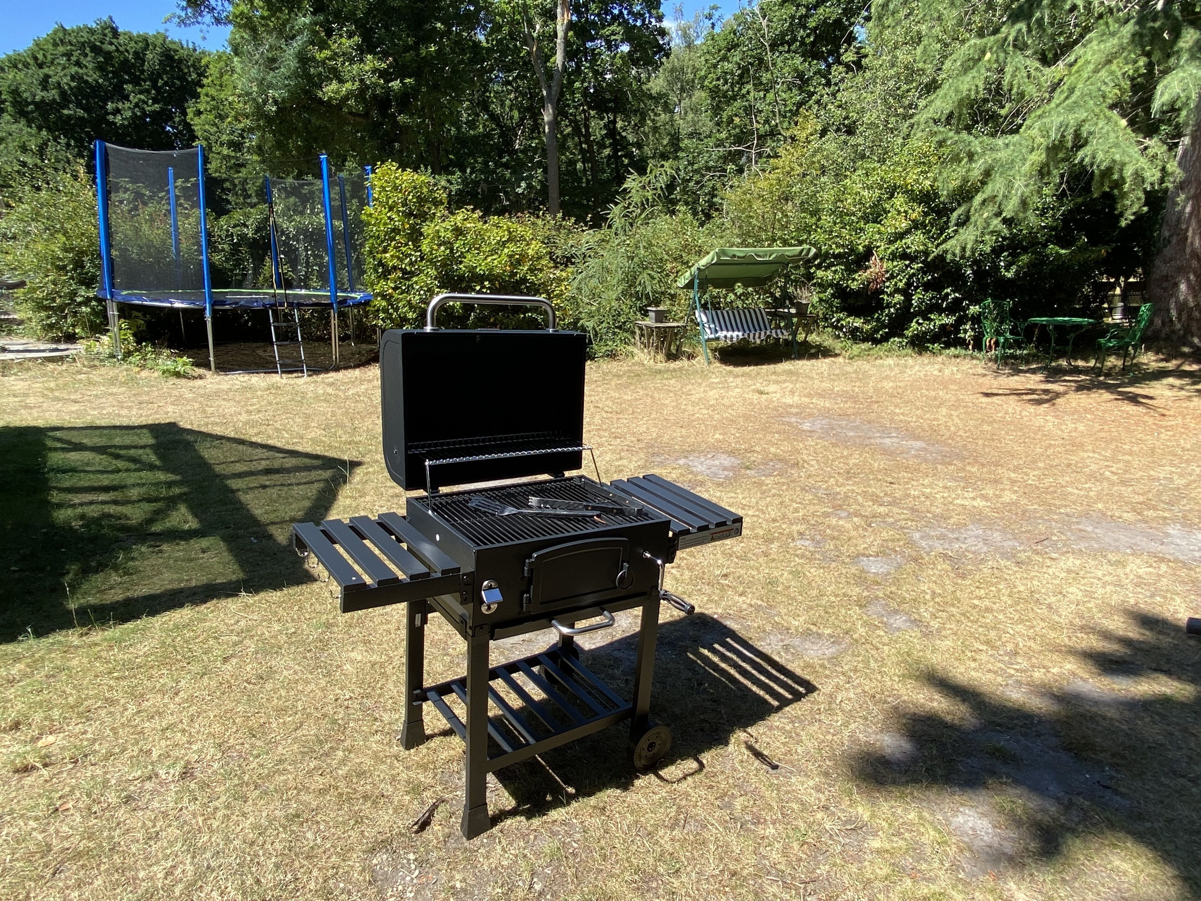 A portable barbecue grill in a grassy outdoor area with trees and a trampoline in the background.