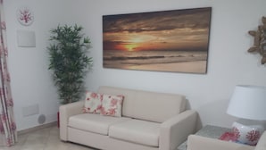 Una sala de estar con un sofá color crema, una planta en maceta, un cuadro colgado en la pared que representa un atardecer y una lámpara sobre una mesita.