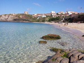 Una playa costera con agua cristalina, afloramientos rocosos y edificios al fondo.