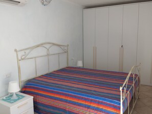 Un dormitorio con estructura de cama metálica, colcha a rayas de colores, mesita de noche con lámpara y armario.