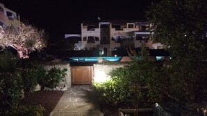 Vista nocturna de un patio de hotel con un camino de piedra que lleva a una puerta de madera, rodeado de vegetación y una zona de piscina.