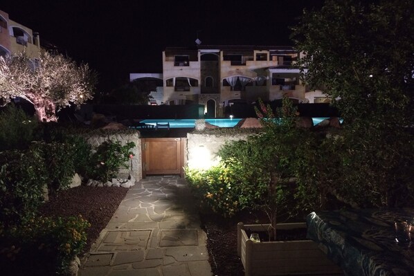 A nighttime view of a hotel courtyard with a stone pathway leading to a wooden door, surrounded by greenery and a pool area.