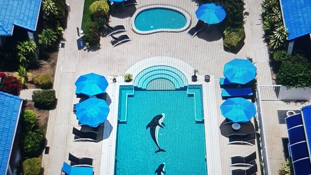 An aerial view of a resort with a central swimming pool, surrounded by lounge chairs and umbrellas, and a building with a red roof in the background. - LARGE VILLA SWIMMING POOLS TROPICAL GARDEN SEA GOLF RELAXATION 6 /12 ADULTS (Phuket)