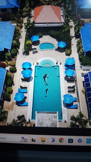 Vista aérea de un resort con una piscina central, rodeada de sillas de descanso y sombrillas, y un edificio con techo rojo al fondo.